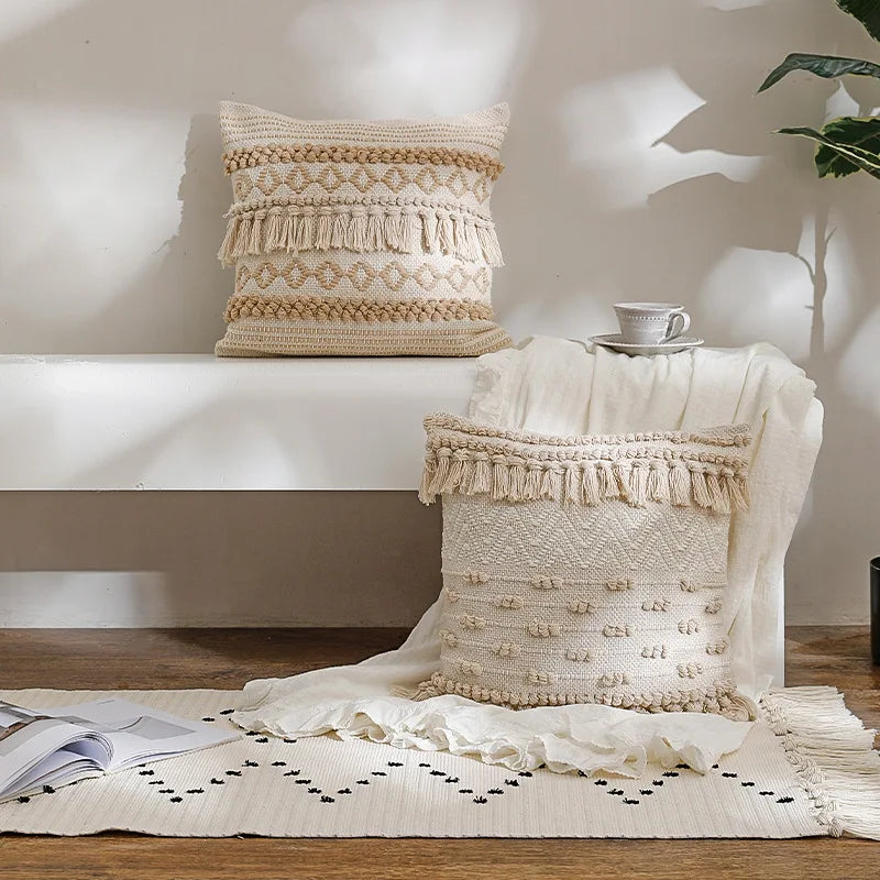 Housse de Coussin Coton Lin 45x45 style bohème avec des motifs tissés en losanges, des franges et des pompons, dans des tons neutres beige et écru, sur un fond blanc - plan large sur banc