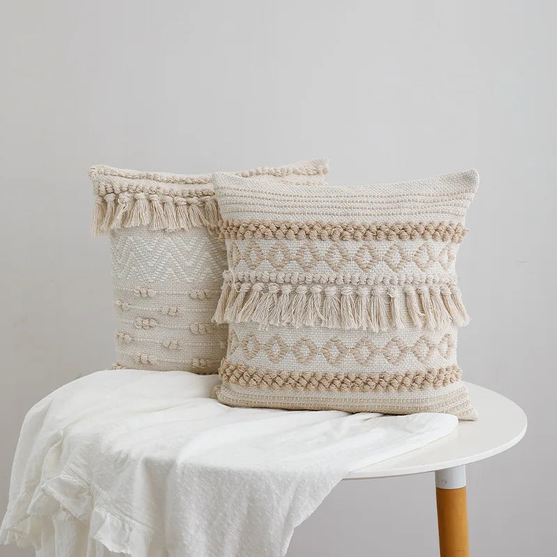 Housse de Coussin Coton Lin 45x45 style bohème avec des motifs tissés en losanges, des franges et des pompons, dans des tons neutres beige et écru, sur un fond blanc - sur table