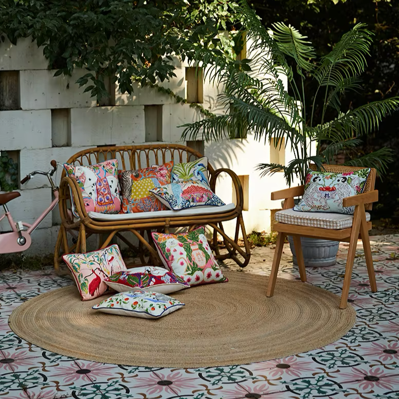 Housse de Coussin Rectangulaire Rose avec bordure rouge vif, décoré de motifs naturalistes montrant des oiseaux exotiques perchés sur des branches d'arbre avec feuilles vertes et fruits jaunes, sur un fond beige rosé - au pied du fauteuil en rotin, plan large sur la scène