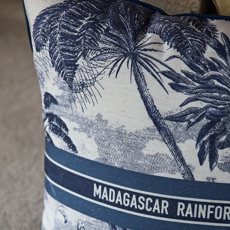 Housse de Coussin 45x45 avec Lettre représentant la forêt tropicale de Madagascar avec des palmiers, des flamants roses et un grand papillon. Le texte "MADAGASCAR RAINFOREST" est centré, encadré par deux lignes horizontales - zoom sur matière