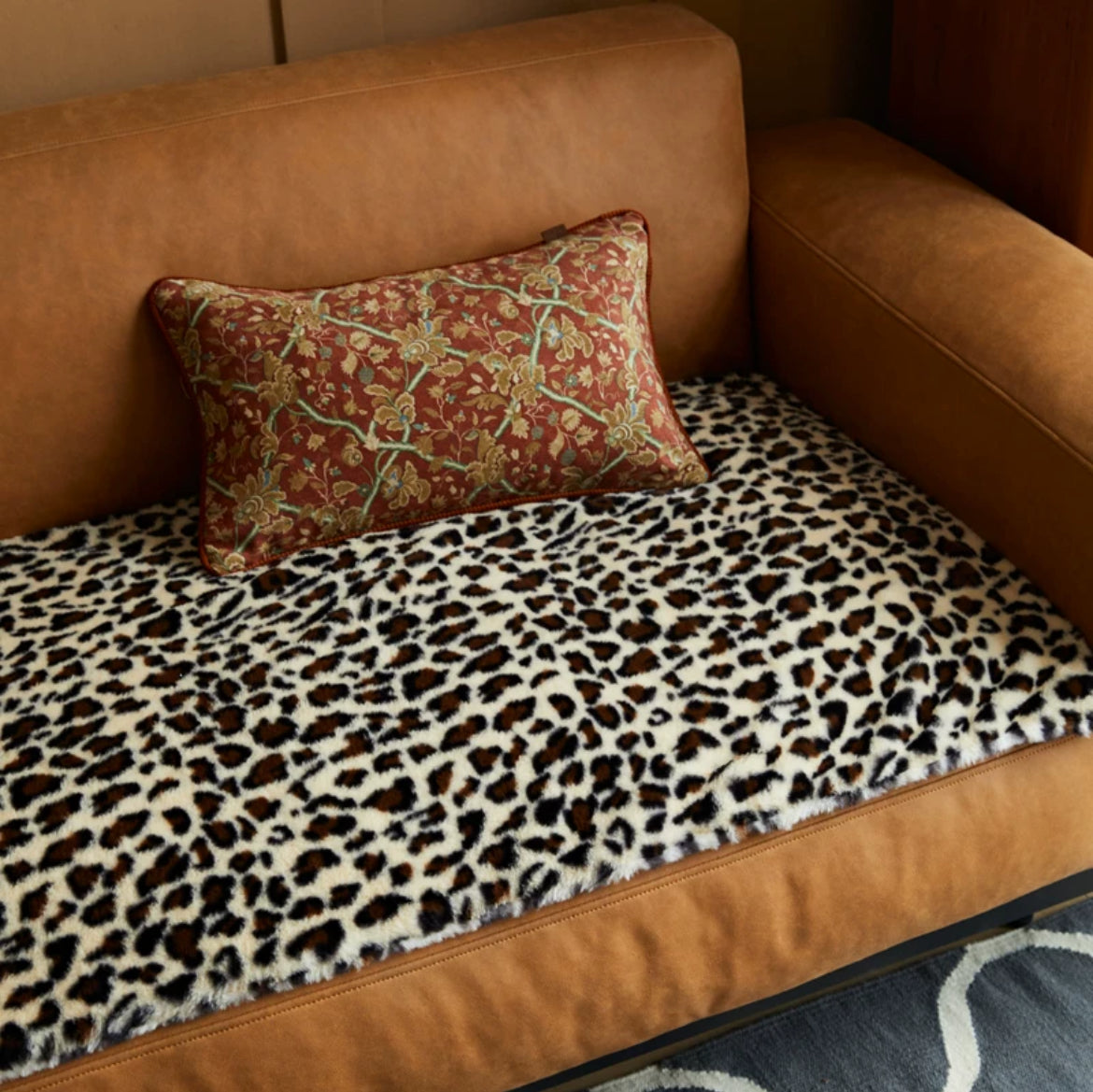 Housse de Coussin Fleurie vu de dessus avec broderies florales dorées et vertes sur un fond rouge brique. Les motifs sont détaillés, les bords arrondis, et une finition en cordon rouge-brun encadre le coussin. Fond blanc pour mettre en valeur le design - version rectangle sur canapé cuir camel