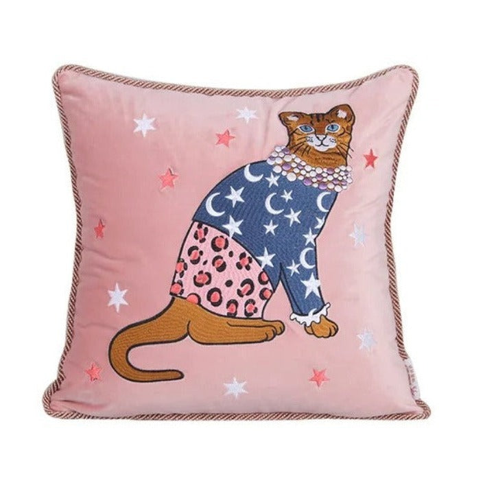 Coussin décoratif avec illustration fantaisiste d'un chat vêtu d'un pull bleu marine étoilé et d'une jupe rose à motif léopard, sur fond rose décoré d'étoiles colorées, avec une bordure torsadée beige et brune.- de face