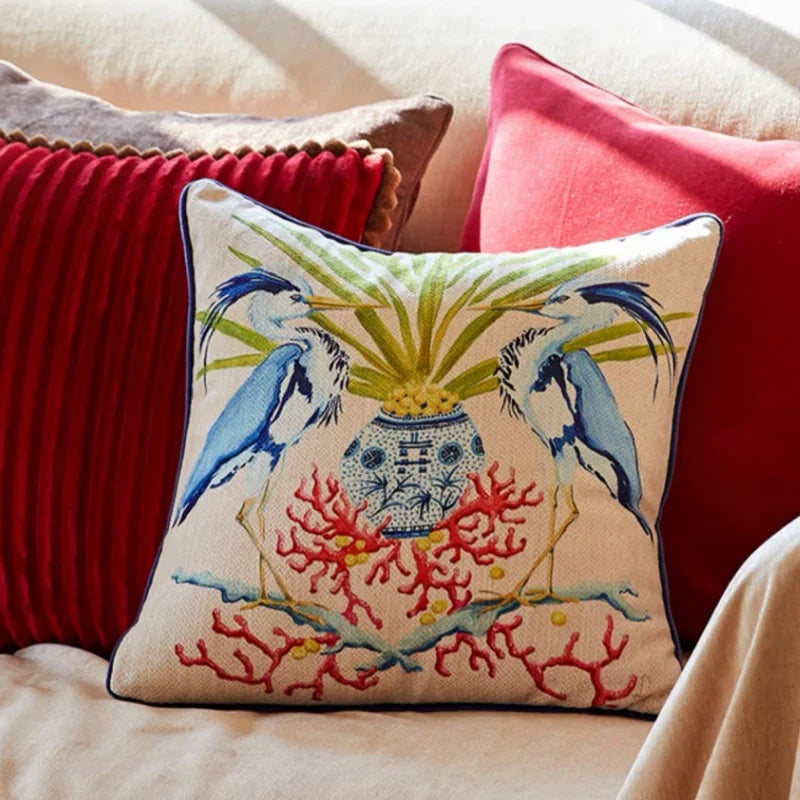 Housse de Coussin Blanc et Bleu décoratif avec deux hérons bleus, un vase orné au centre, des feuilles tropicales vertes et des coraux rouges, style aquarelle tropical et marin - sur canapé