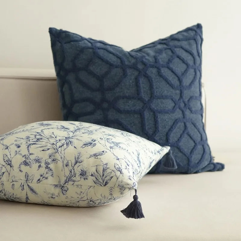 Housse de Coussin 45x45 Lin Blanc décoratif carré avec motifs floraux bleus sur fond blanc, agrémenté de pompons bleus aux quatre coins, présenté sur un arrière-plan clair et minimaliste - version carré couchée sur canapé
