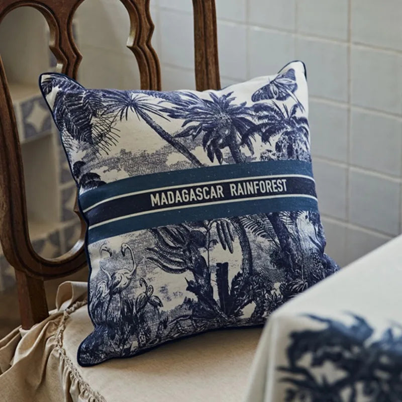 Housse de Coussin 45x45 avec Lettre représentant la forêt tropicale de Madagascar avec des palmiers, des flamants roses et un grand papillon. Le texte "MADAGASCAR RAINFOREST" est centré, encadré par deux lignes horizontales - mis en scène sur chaise