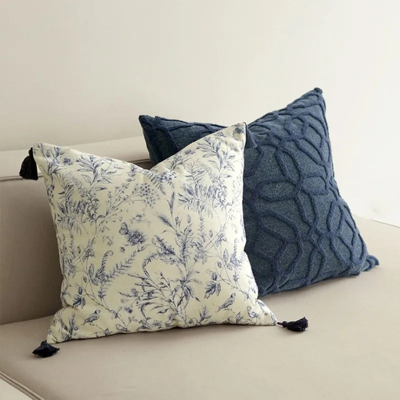 Housse de Coussin 45x45 Lin Blanc décoratif carré avec motifs floraux bleus sur fond blanc, agrémenté de pompons bleus aux quatre coins, présenté sur un arrière-plan clair et minimaliste - sur canapé 