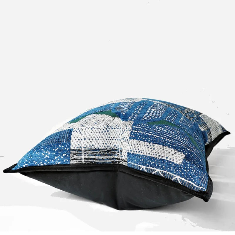 housse de coussin bleu et argent contemporain, avec un design abstrait tissé en bleu, blanc et vert, souligné par un passepoil noir. Fond uni blanc, idéal pour une présentation de produit - couché