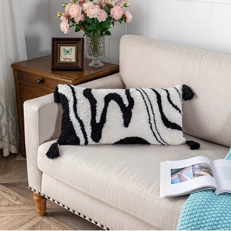 Housse de Coussin Rectangulaire en Noir et Blanc  avec texture bouclée, motif graphique de lignes abstraites et pompons noirs aux coins, sur fond blanc épuré - deuxième photo sur canapé